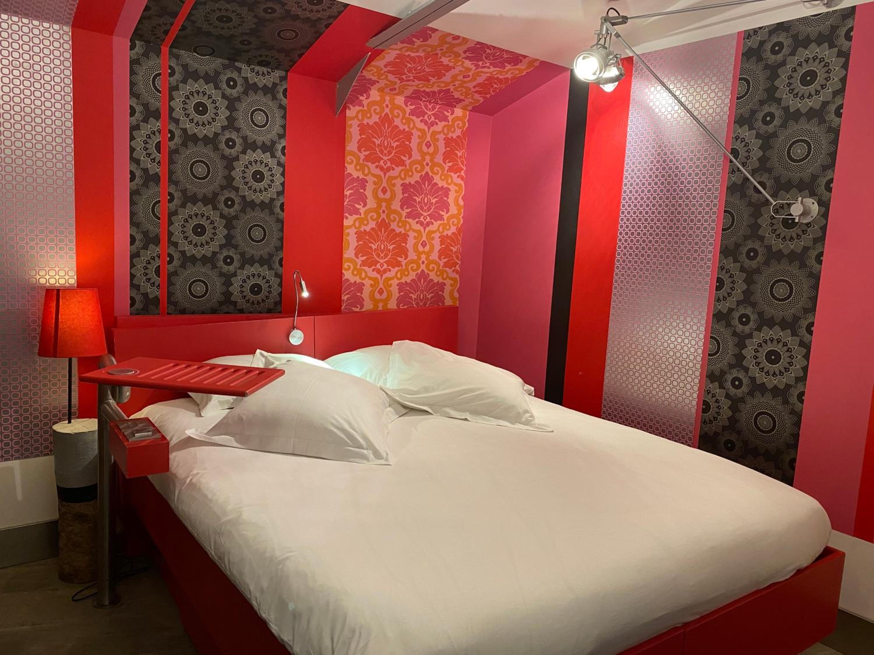 Una cama con ropa blanca, una mesita de noche roja y una lámpara.