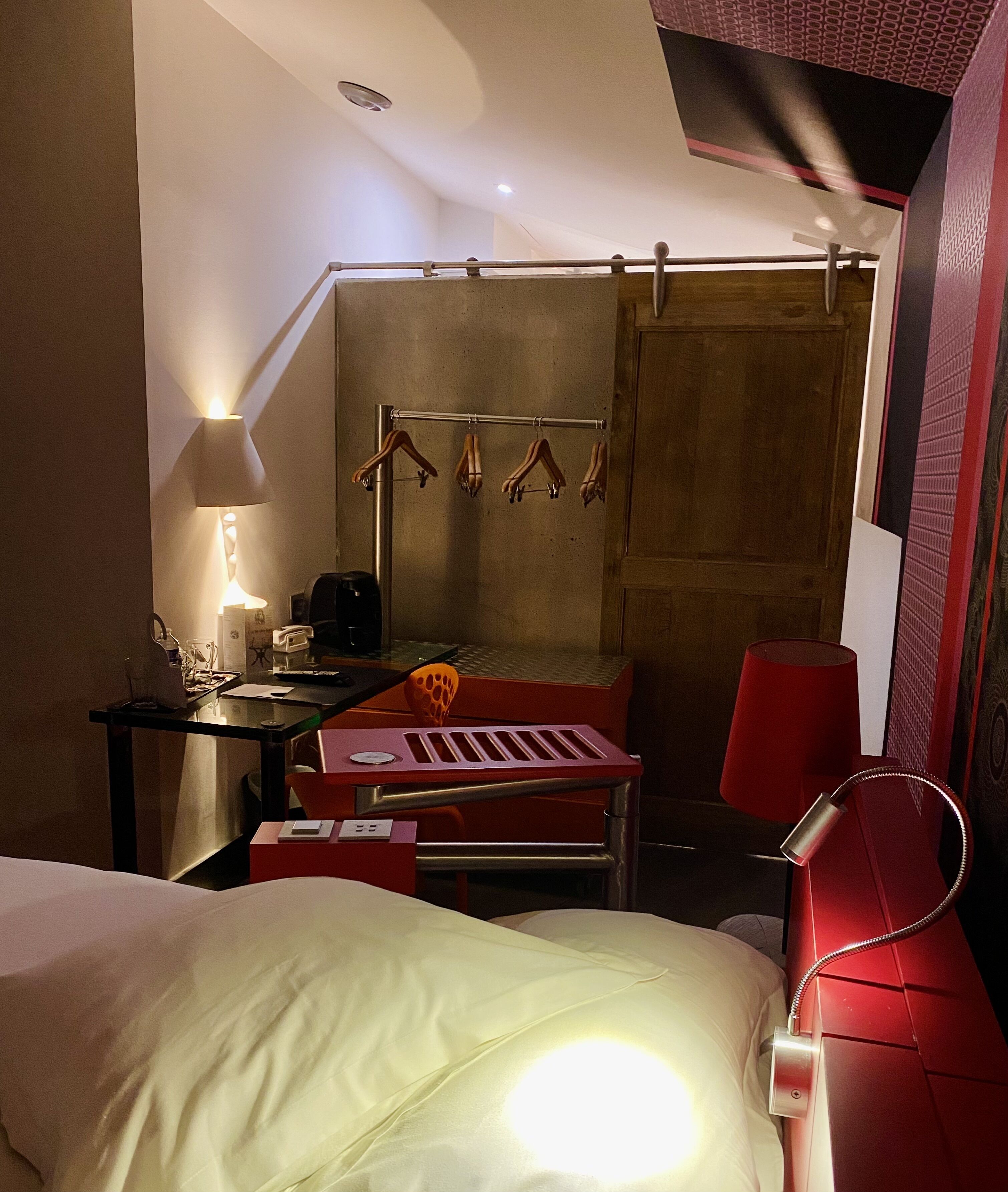 Une chambre d’hôtel de taille modeste, comprenant un lit, un bureau avec une lampe, une chaise, un canapé rouge et un porte-manteau avec des cintres.