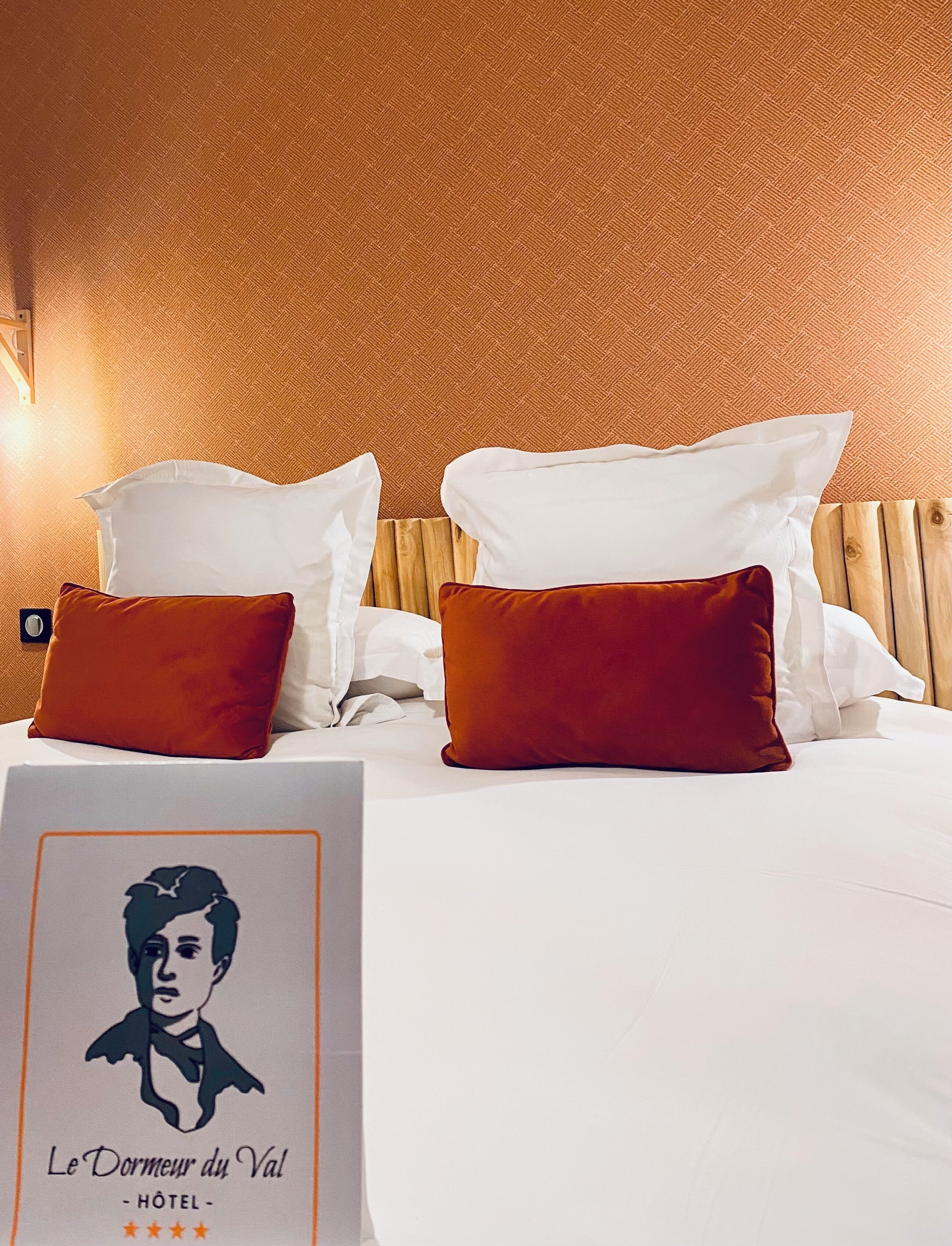 A hotel room with a wooden headboard, white bedding, and two red pillows. A sign with a portrait and text is visible on the bed.