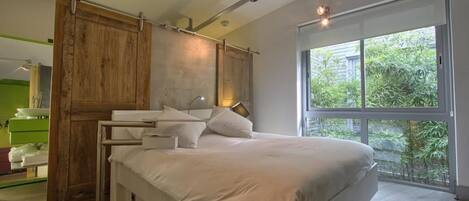 A modern bedroom with a large bed, wooden paneling, a ceiling fan, and a large window with a view of greenery.