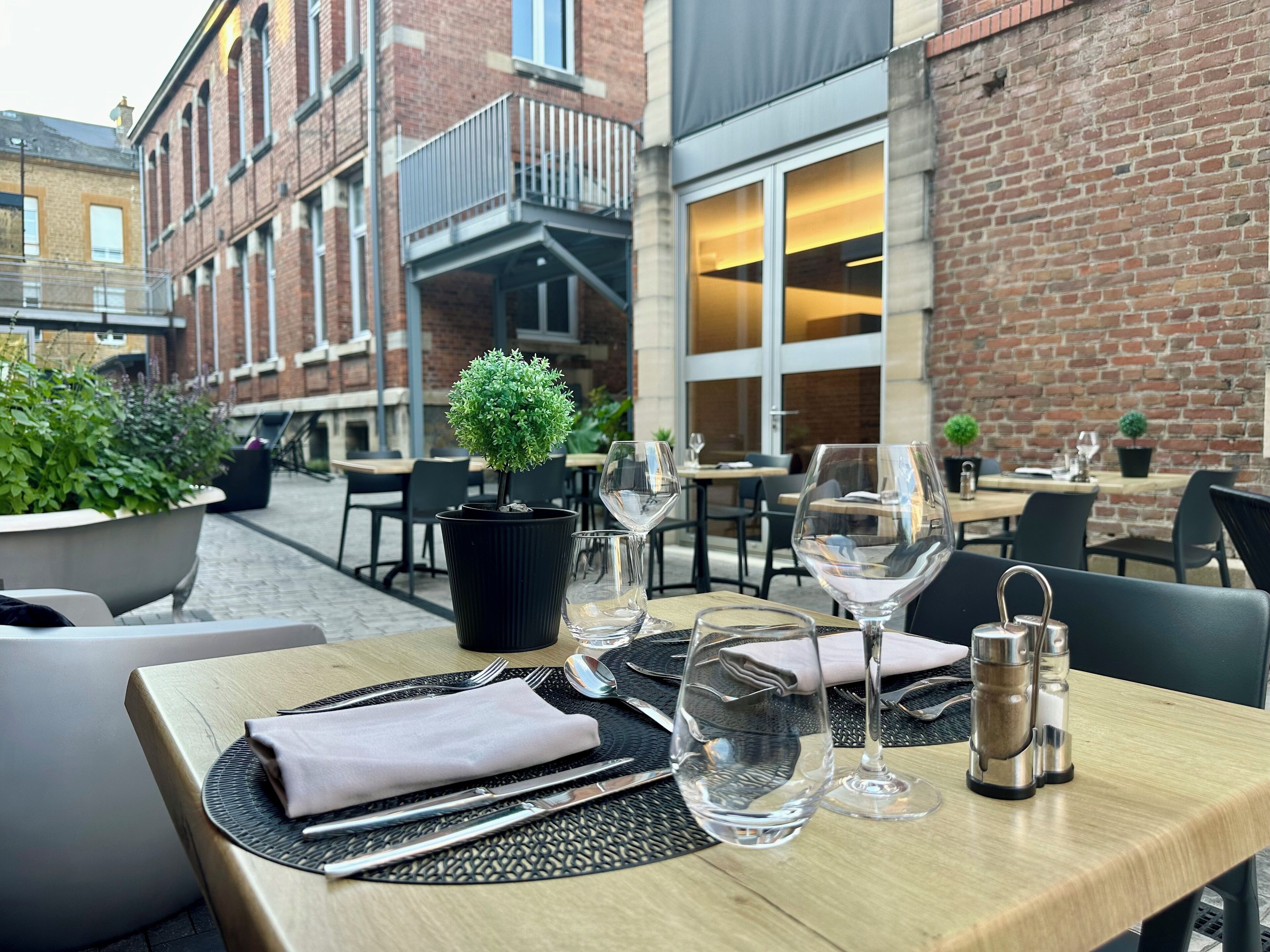 An outdoor dining area with tables set for two, potted plants, and a brick building in the background.
