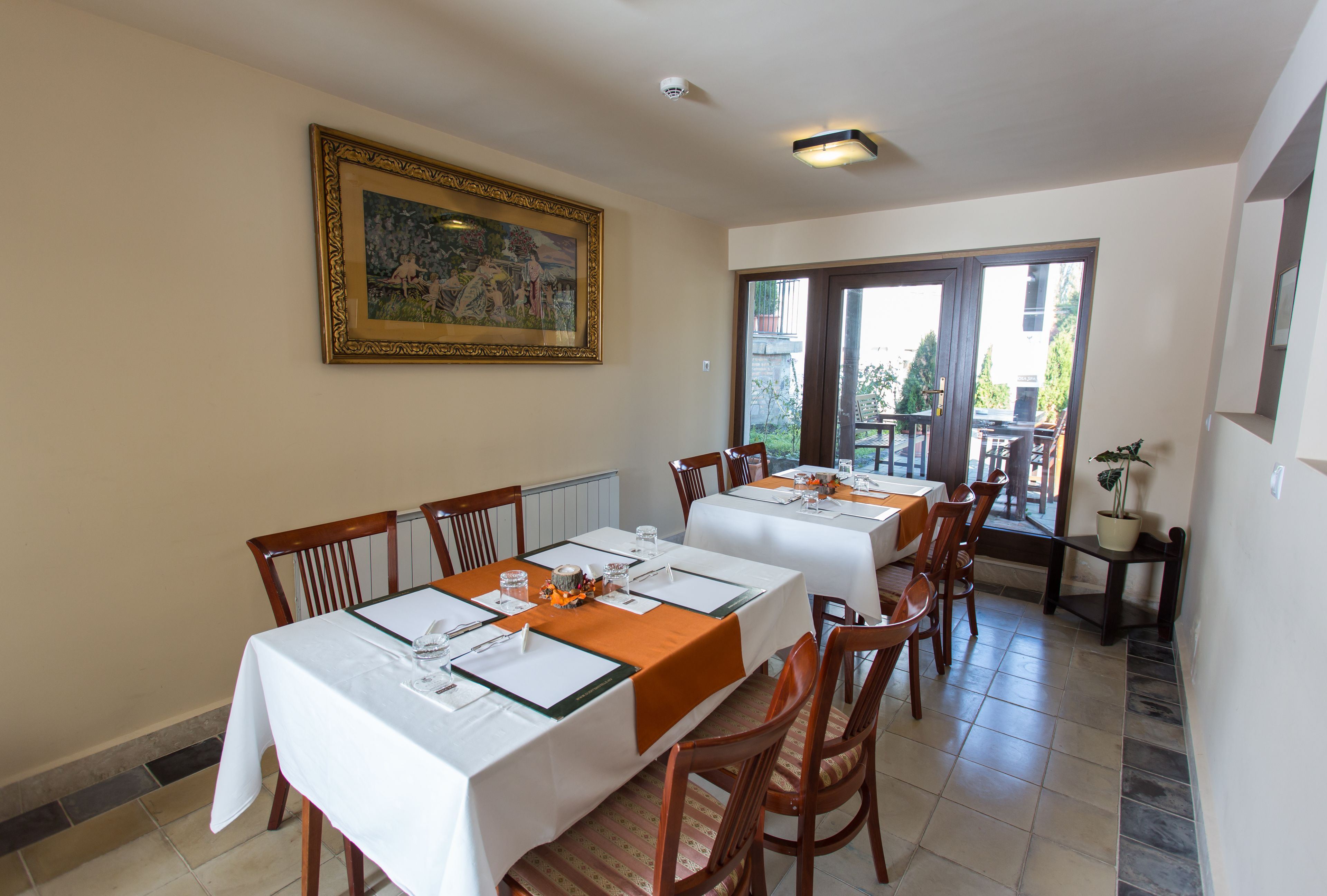 Un comedor con mesas dispuestas para cuatro, sillas, un cuadro grande enmarco y vistas a través de puertas de cristal.