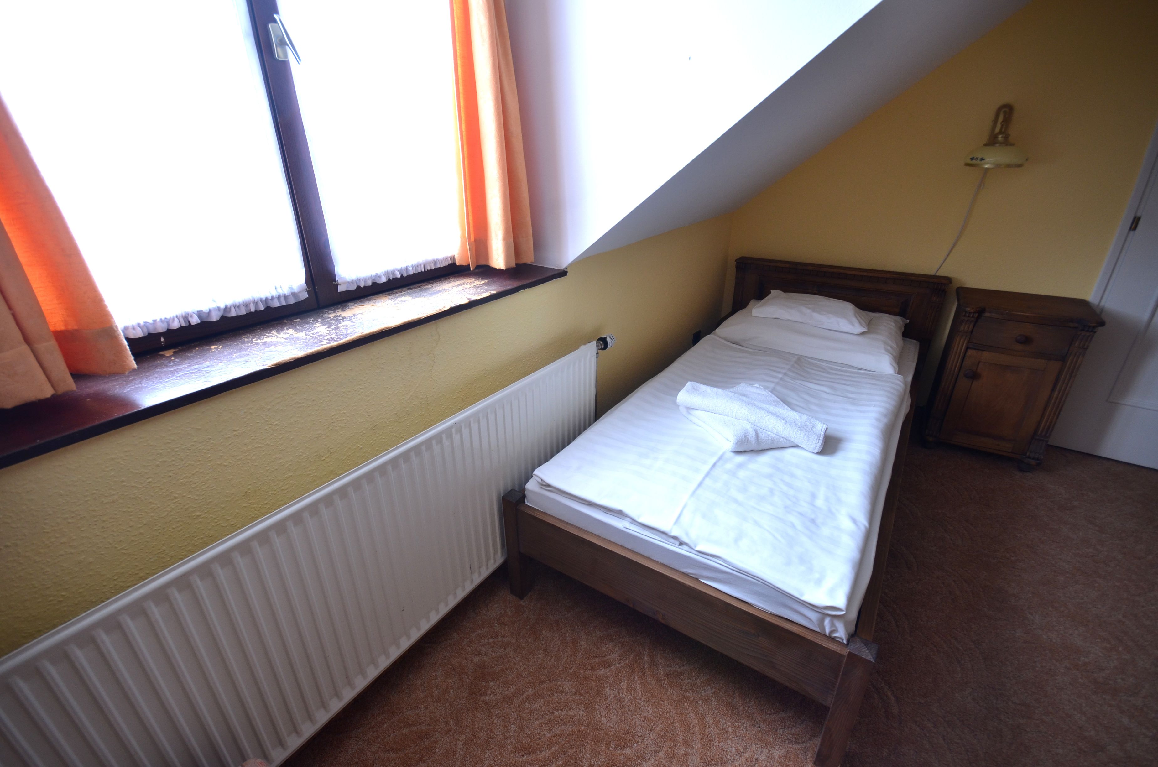 Una habitación pequeña con cama individual, techo inclinado, radiador, mesita de noche de madera y ventana con cortinas.