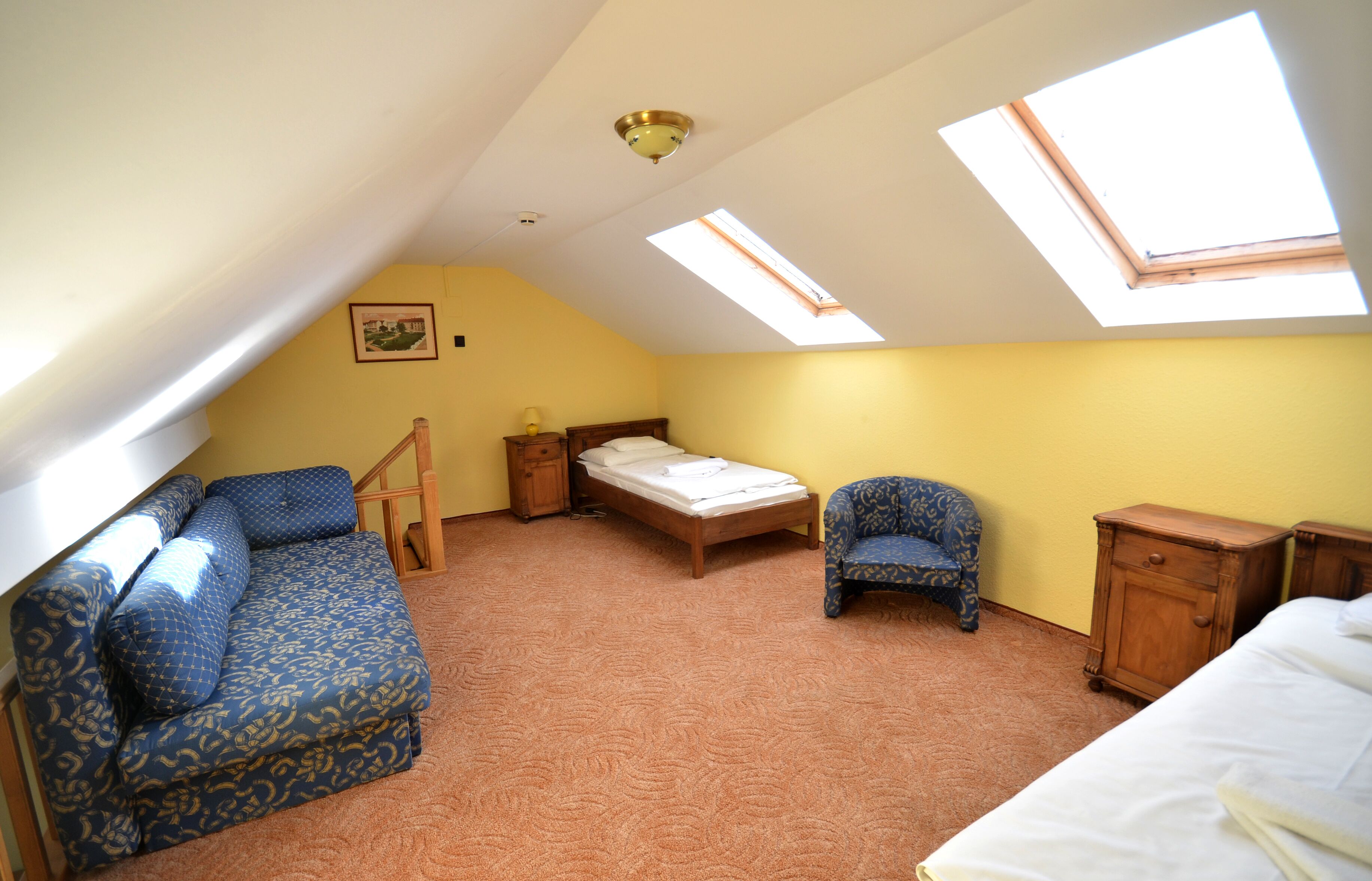 A bedroom with a sloped ceiling, a bed, a sofa, a chair, and a small cabinet.