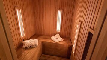 A sauna with wooden walls and benches, two small windows with light fixtures, and a towel on one bench.