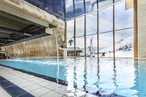 An indoor swimming pool with a waterfall feature, large windows, and a view of snow-capped mountains. - Hotel Edelweiss (Soelden)