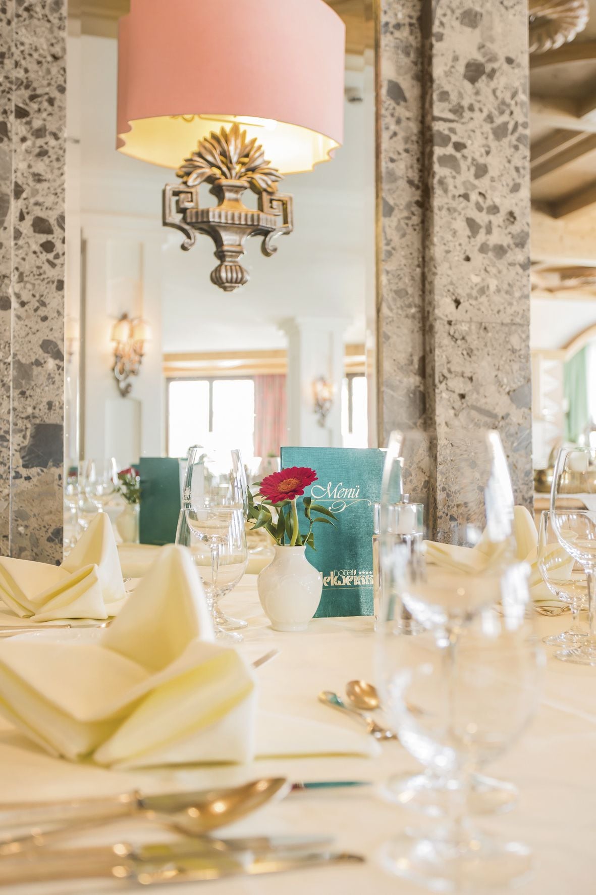 Une table à manger dressée avec des serviettes blanches, des verres à vin et un vase contenant une fleur rouge.