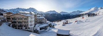 Estância de montanha coberta de neve, com vários edifícios, incluindo um hotel com varandas e um teleférico ao fundo.