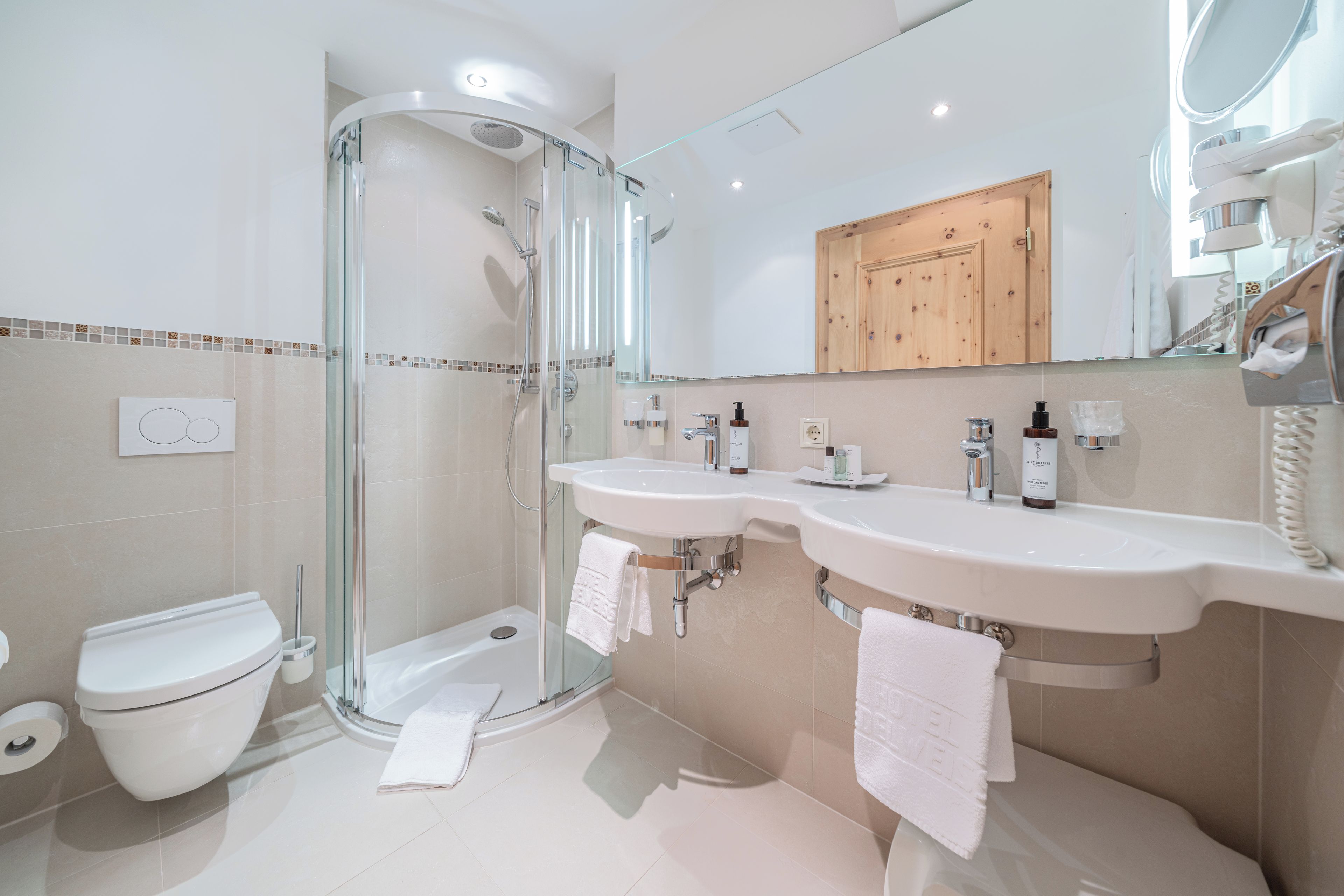 Een moderne badkamer met een douche met glazen wand, twee wastafels, een toilet en een houten kastje boven de spiegels.
