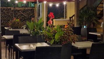 Un área de comedor al aire libre con mesas y sillas, rodeada de plantas en macetas y luces colgantes.