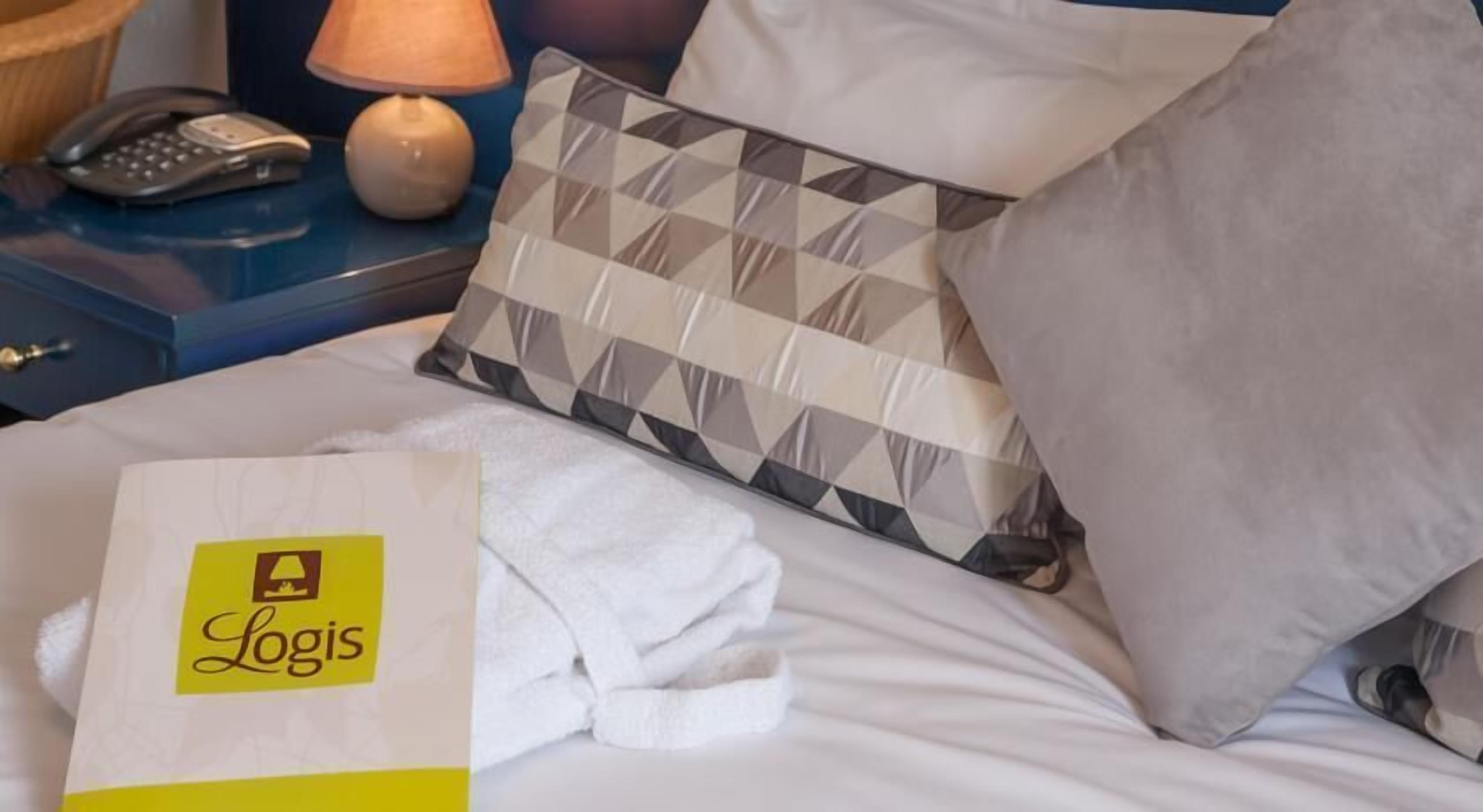 Ein ordentlich bezogenes Bett mit einem weißen Handtuch, einem gemusterten Kopfkissen und einer weißen Tasche mit einem Logo.