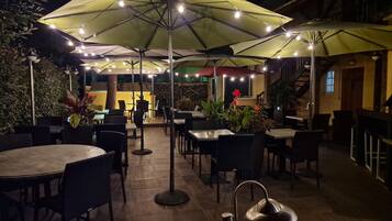 Een buitenterras met tafels en stoelen, afgedekt door parasols en lichtslingers.