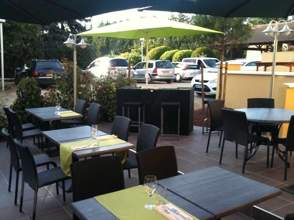 Une terrasse extérieure avec des tables et des chaises, un espace salon couvert et, en arrière-plan, une vue sur les voitures garées.