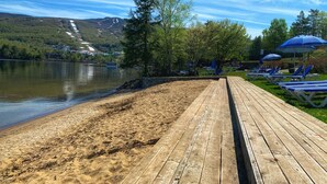 Un muelle de madera que lleva a una playa arenosa con sillones de playa y sombrillas, con vistas a un lago tranquilo y un paisaje montañoso.