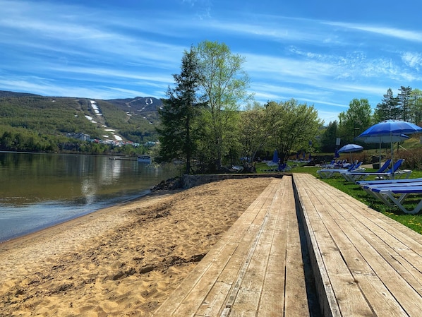 A wooden pier leading to a sandy beach with lounge chairs and umbrellas, overlooking a calm lake and a mountainous landscape.