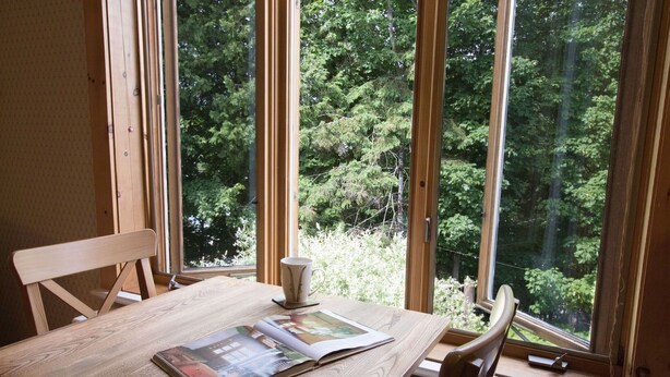 Una mesa de madera con una revista, una taza y una silla en una habitación con ventanas grandes que dan a un bosque.