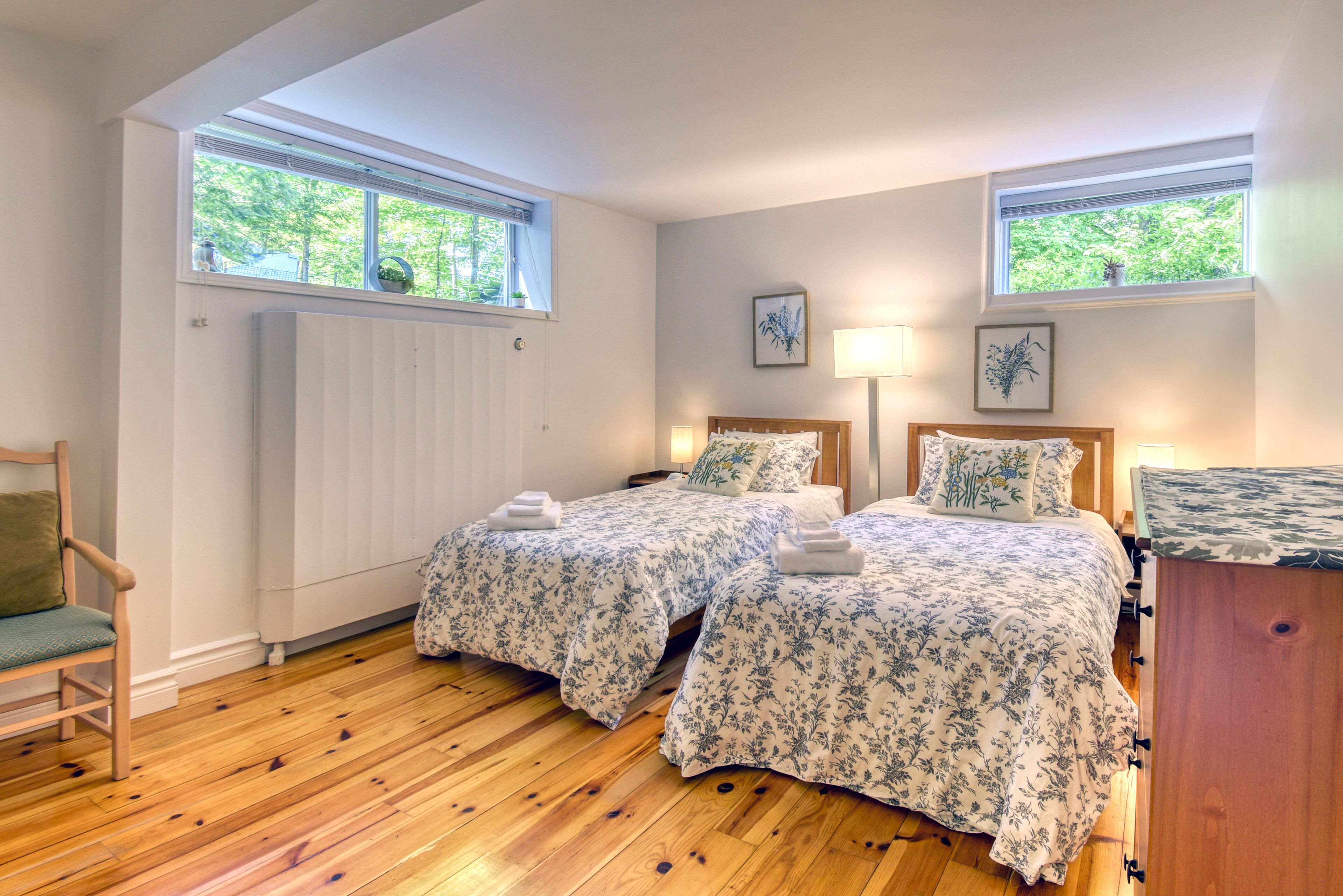 Une chambre avec deux lits, un plancher en bois, une chaise et deux fenêtres équipées de stores.