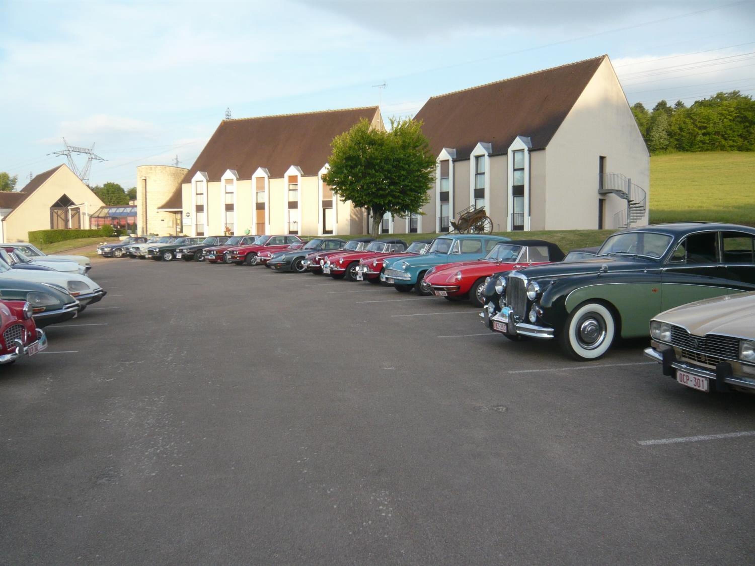 A parking lot with vintage cars lined up in front of a row of buildings with sloped roofs.