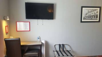 A hotel room with a flat-screen TV mounted on the wall, a desk with a chair, a radiator, a water bottle, and a framed picture.