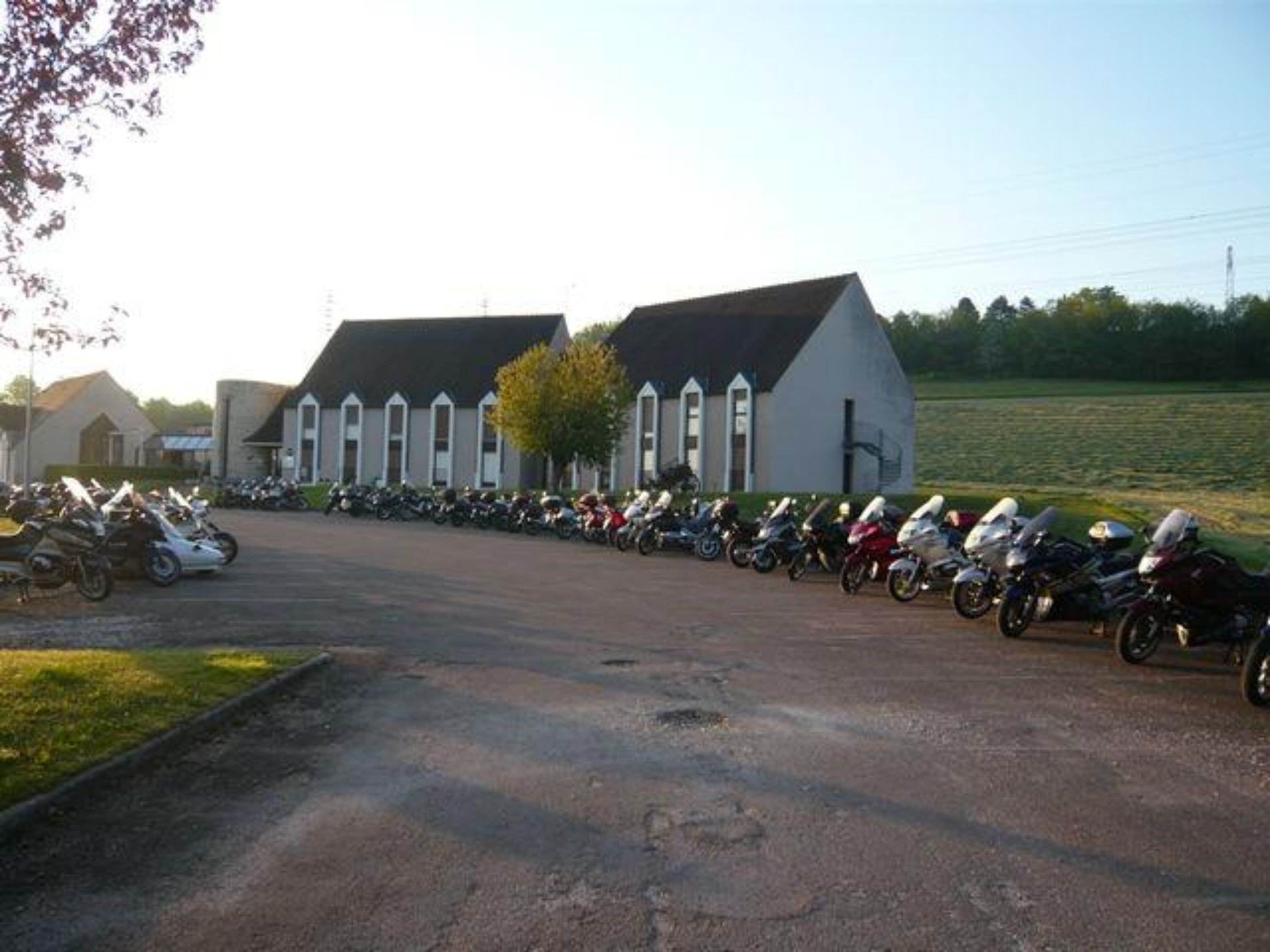 A parking lot with multiple motorcycles lined up in front of a building with a distinct architectural style, including pointed windows and a steeply pitched roof.