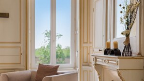 Une pièce avec un fauteuil beige, une cheminée blanche agrémentée d’objets décoratifs et une vue dégagée sur l’extérieur grâce à une grande fenêtre.