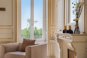 Um quarto com uma poltrona bege, uma lareira branca com objetos decorativos e vista através de uma grande janela.