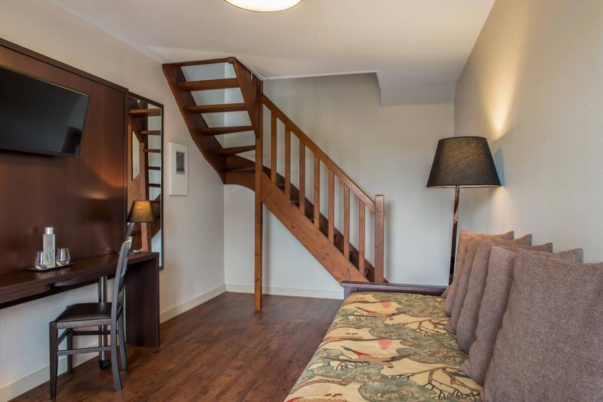 Una sala de estar con una escalera de madera, un sofá con cojines estampados, un televisor montado en la pared y una lámpara sobre una mesita auxiliar.