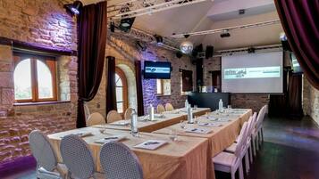 A conference room with long tables, chairs, a projector screen, and a stone wall.