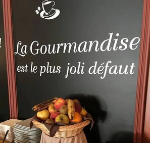 Un mur sur lequel est inscrit le texte « La gourmandise est le plus joli des défauts » et un panier de fruits.
