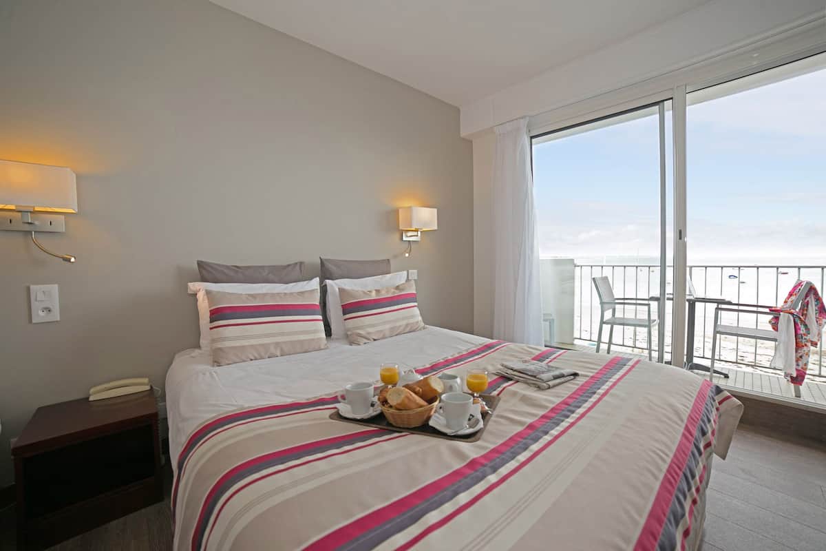 Habitación de hotel con una cama grande, dos lámparas de noche, una mesita de noche y un balcón con vistas a la playa.