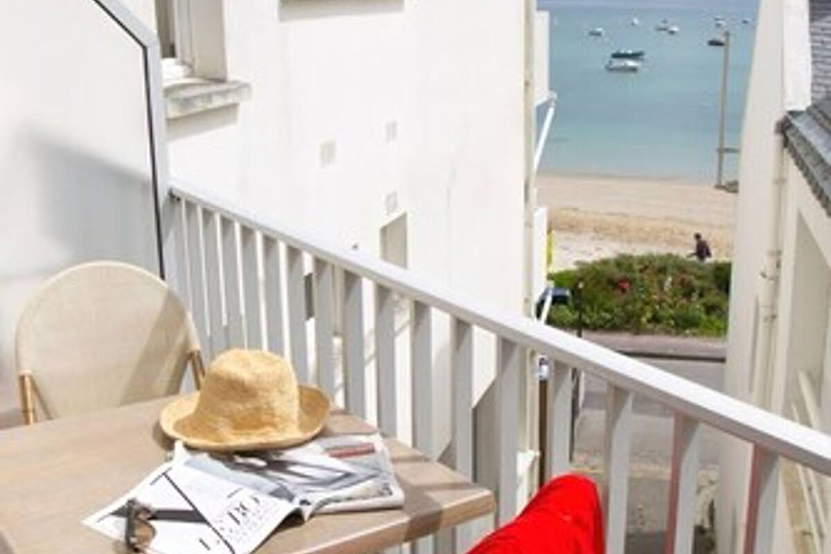 Un balcón con una mesa, sillas y un paño rojo. Hay un sombrero de paja sobre la mesa. Se ve una playa y barcos.