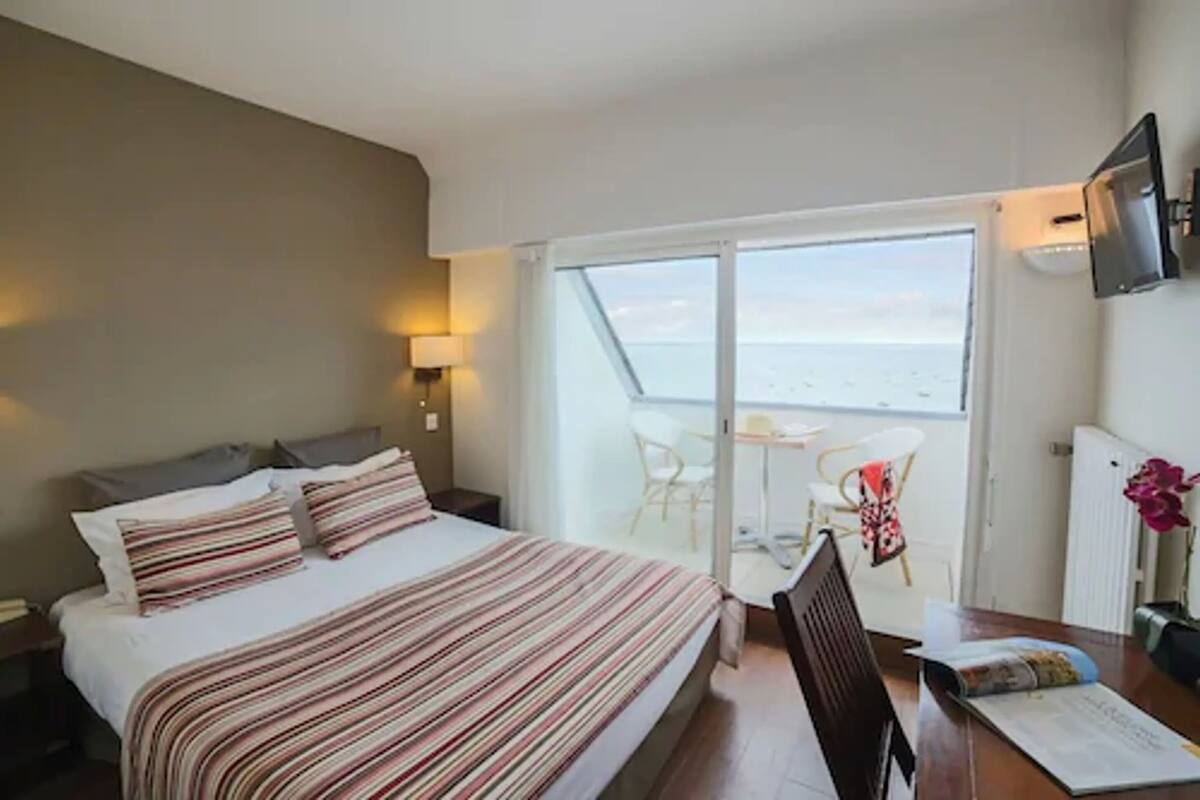 Habitación de hotel con cama, escritorio, silla y vista al mar a través de un ventanal.