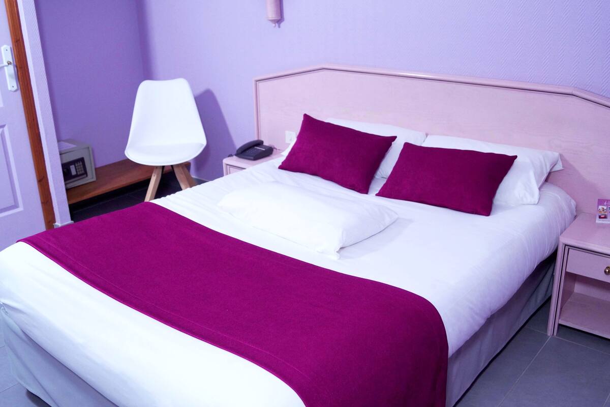 Una cama bien hecha con ropa de cama blanca y morada, una mesita de noche con una lámpara y otra mesita de noche pequeña con un teléfono.