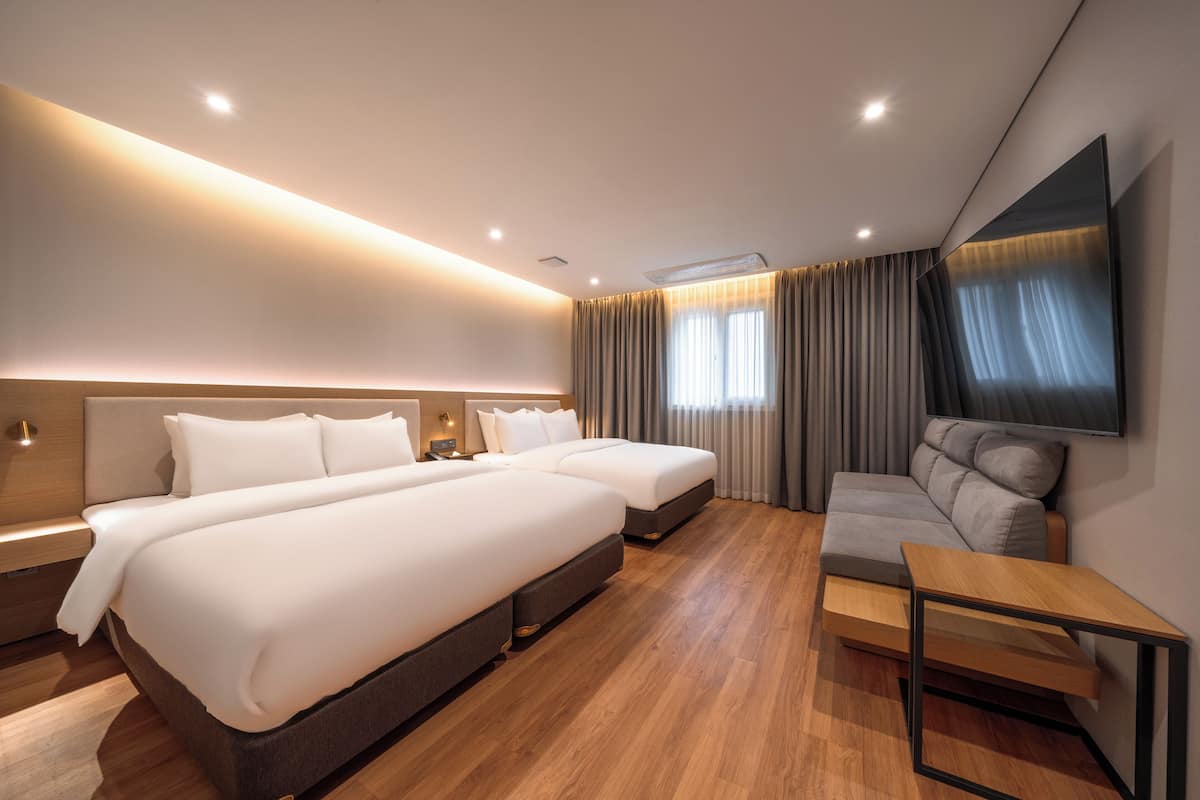 Una habitación de hotel moderna con dos camas, un sofá, un televisor y suelo de madera.