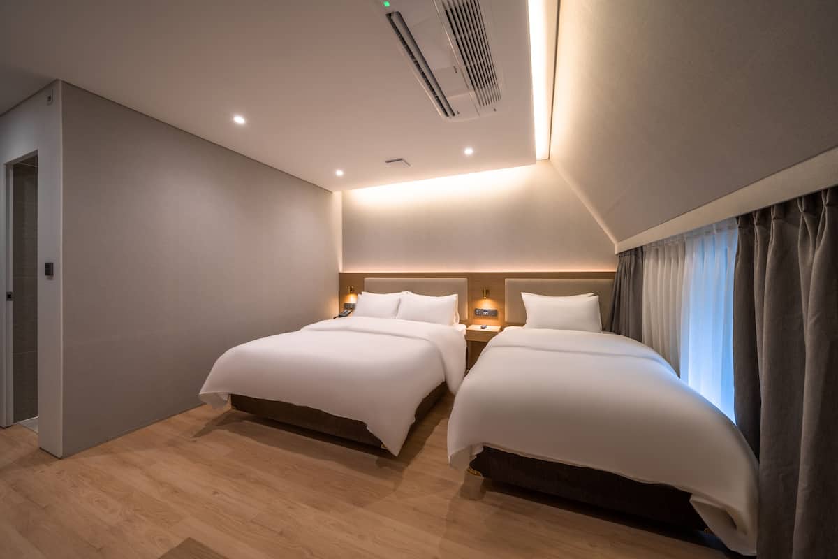 Habitación de hotel con dos camas, piso de madera, un respiradero en el techo y una ventana con cortinas.