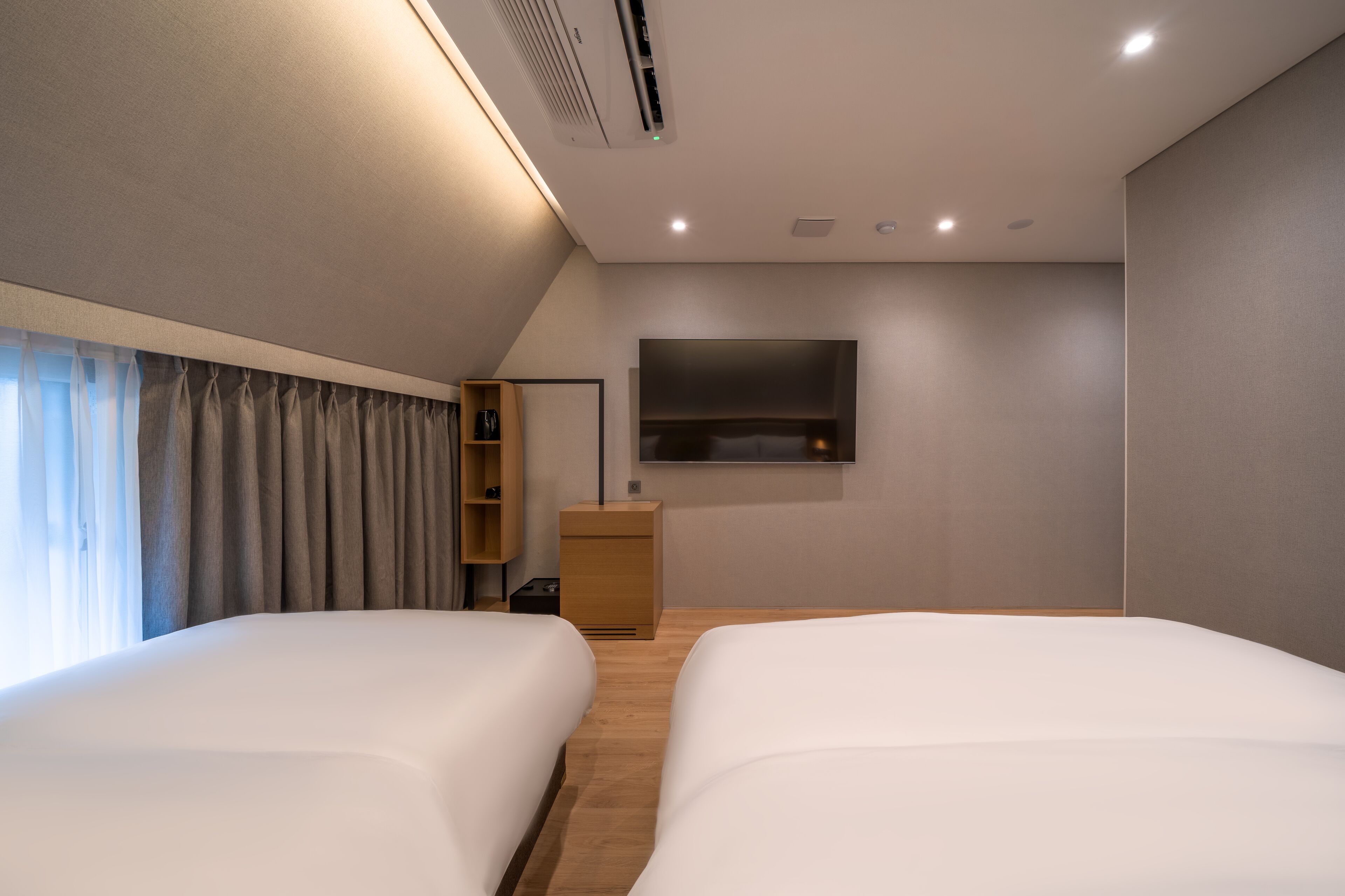 A modern hotel room with two beds, a flat-screen TV mounted on the wall, a wooden nightstand, and a built-in shelf.