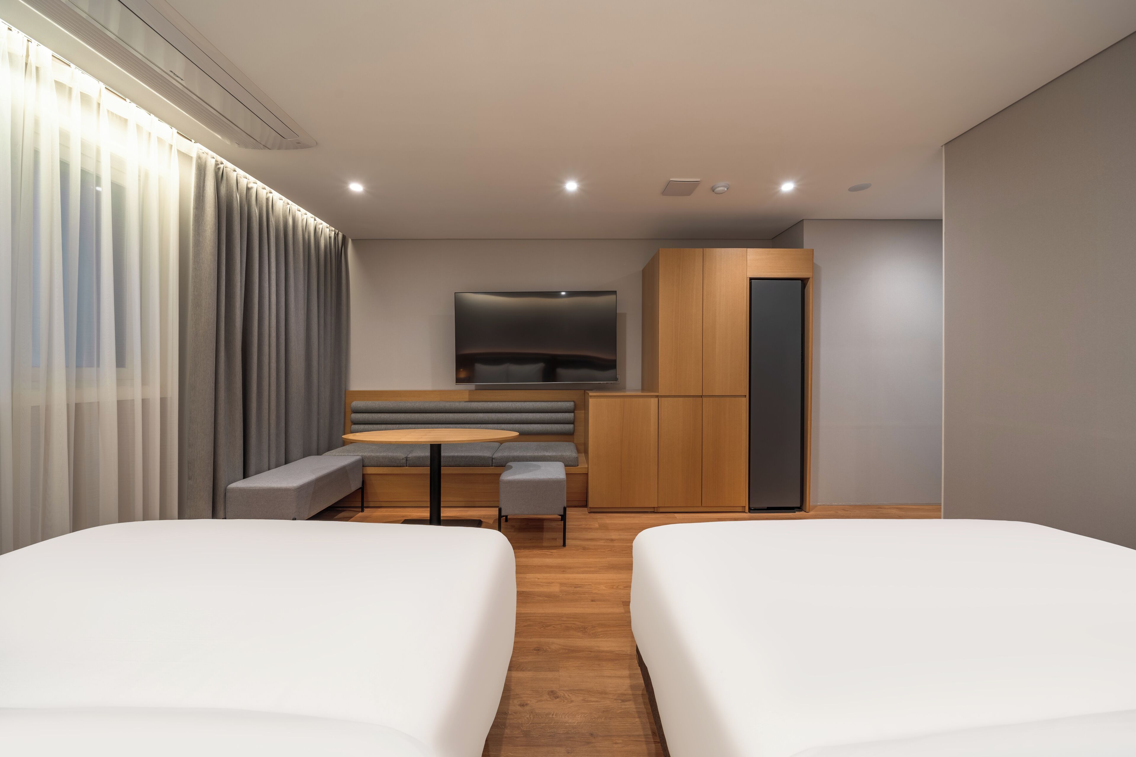 A modern hotel room with two beds, a small seating area, a TV, and a wooden cabinet.