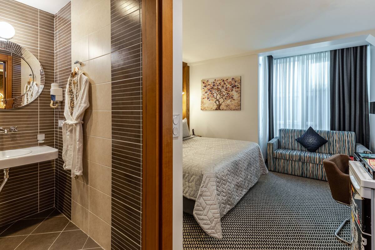 A modern hotel room with a bathroom featuring a sink, mirror, and towel rack. The bedroom has a bed, a sofa, a desk, and a window with curtains.