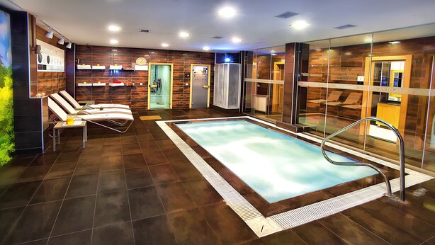 Un área interior de piscina moderna con una piscina rectangular, sillones de descanso y una sauna con paredes de vidrio.