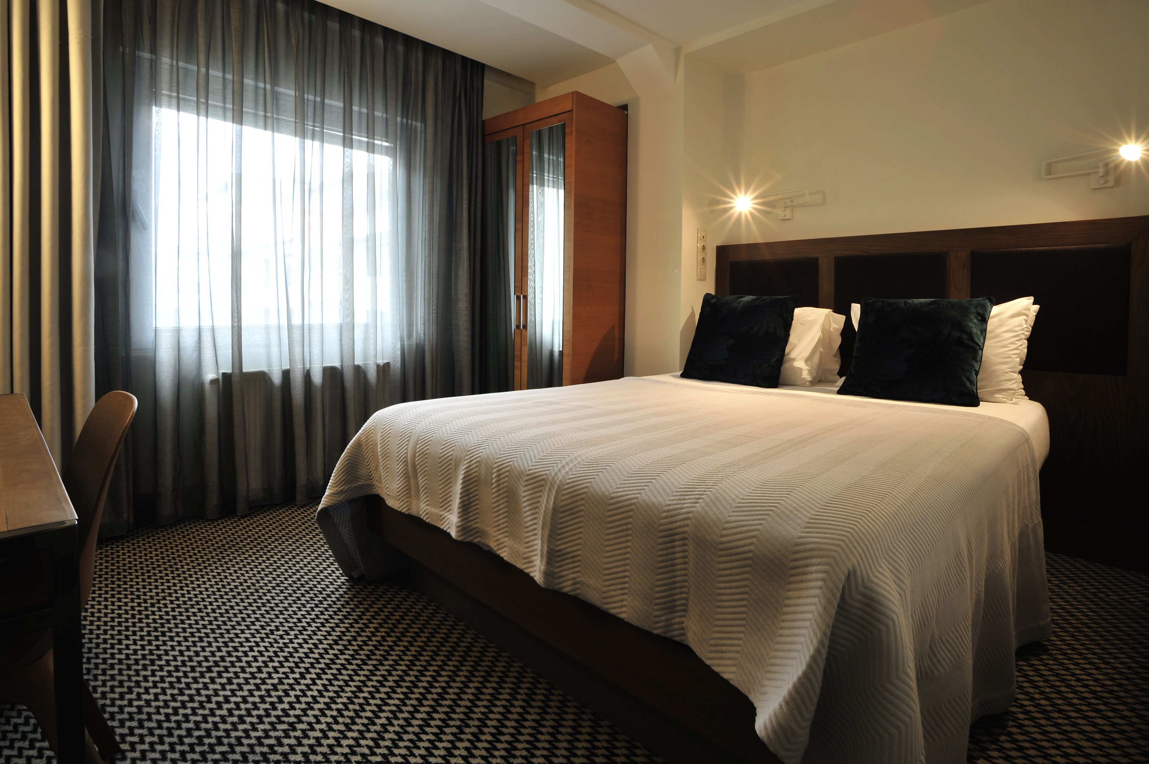 A hotel room with a large bed, a desk, and a chair. There is a window with curtains and a headboard with pillows.