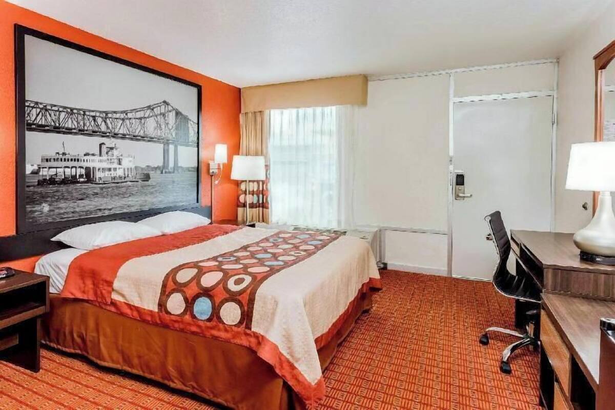 A hotel room with a bed, a desk, a chair, a lamp, a window with curtains, and a painting of a bridge.
