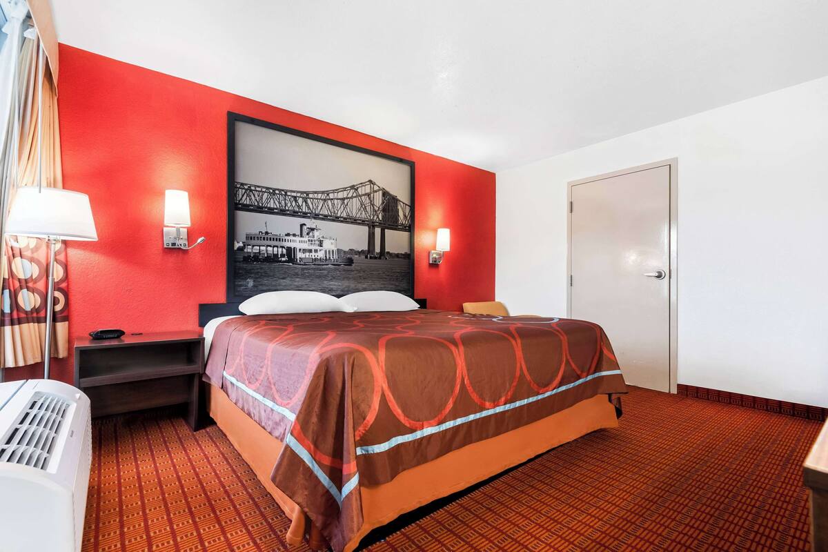 A hotel room with a red wall, a bed with a patterned bedspread, two bedside lamps, a nightstand, a wall-mounted picture of a bridge, and a door.