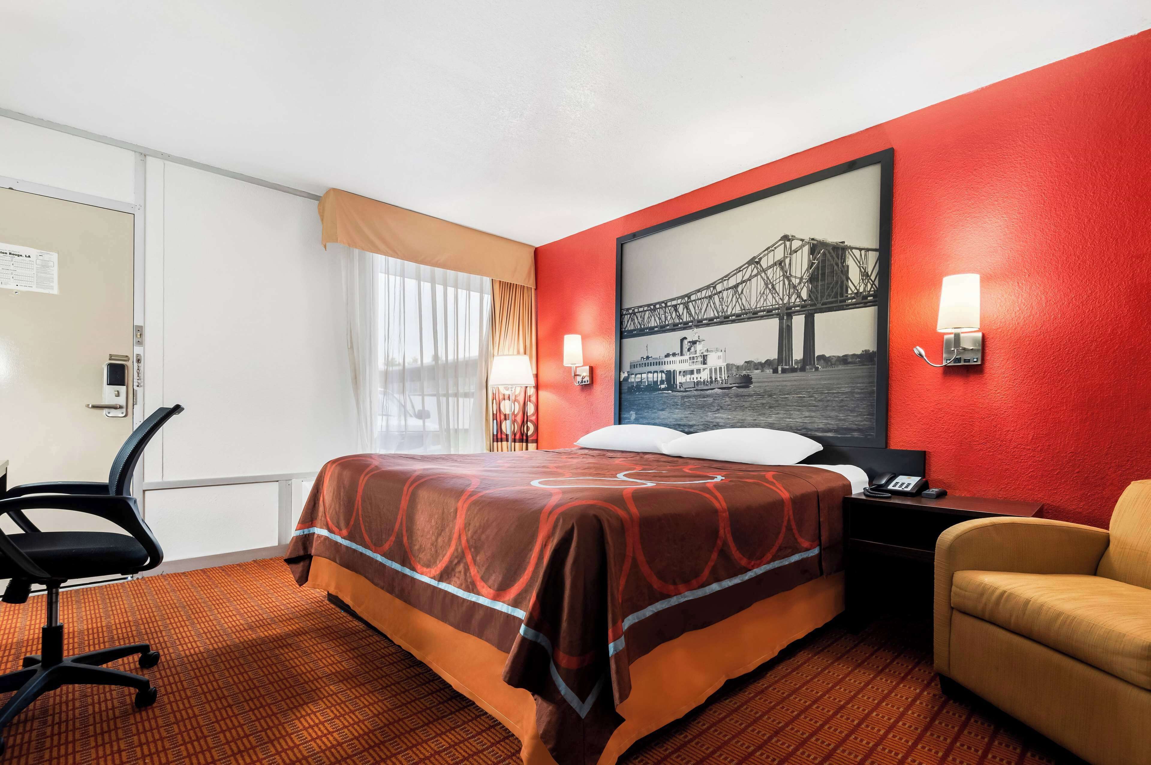 A hotel room with a bed, a desk, a chair, a nightstand, and a wall-mounted lamp.