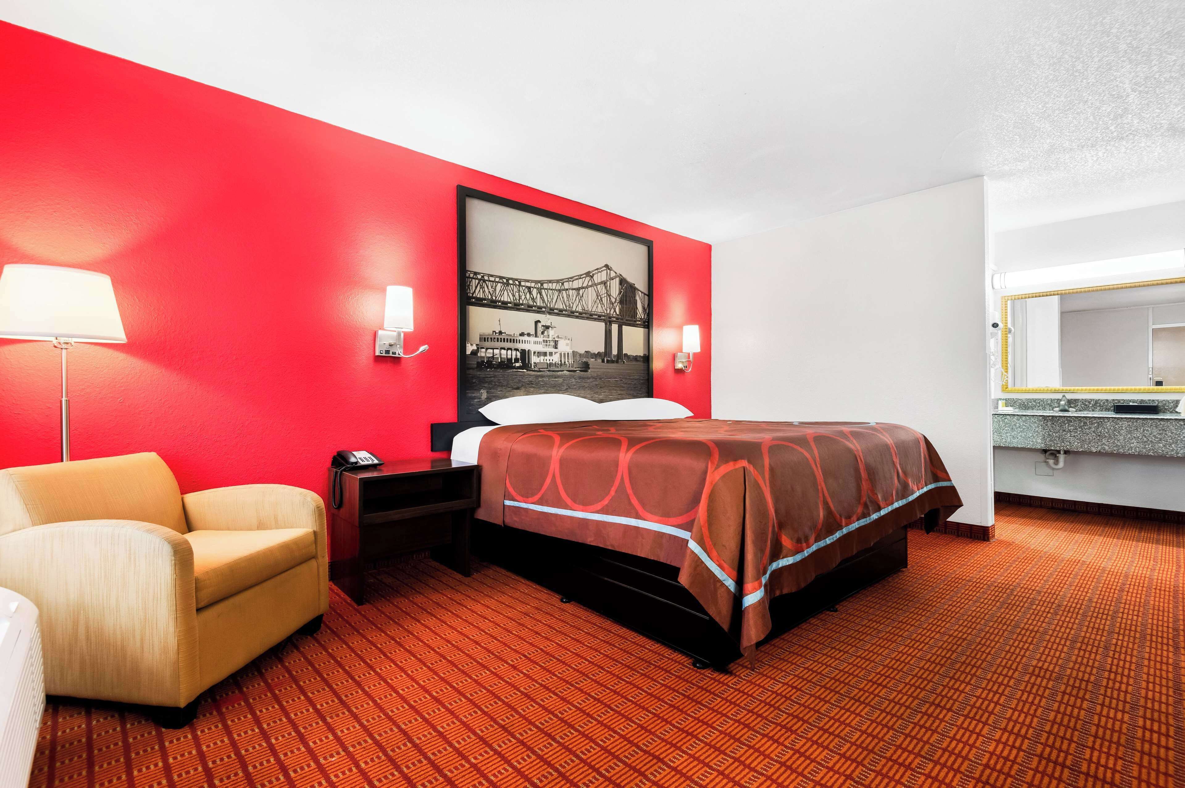 A hotel room with a red accent wall, a bed with a patterned bedspread, a chair, a nightstand, a lamp, and a framed picture of a bridge.