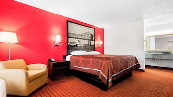 Ein Hotelzimmer mit roter Akzentwand, einem Bett mit gemustertem Überwurf, einem Sessel, einem Nachttisch, einer Lampe und einem gerahmten Bild einer Brücke.