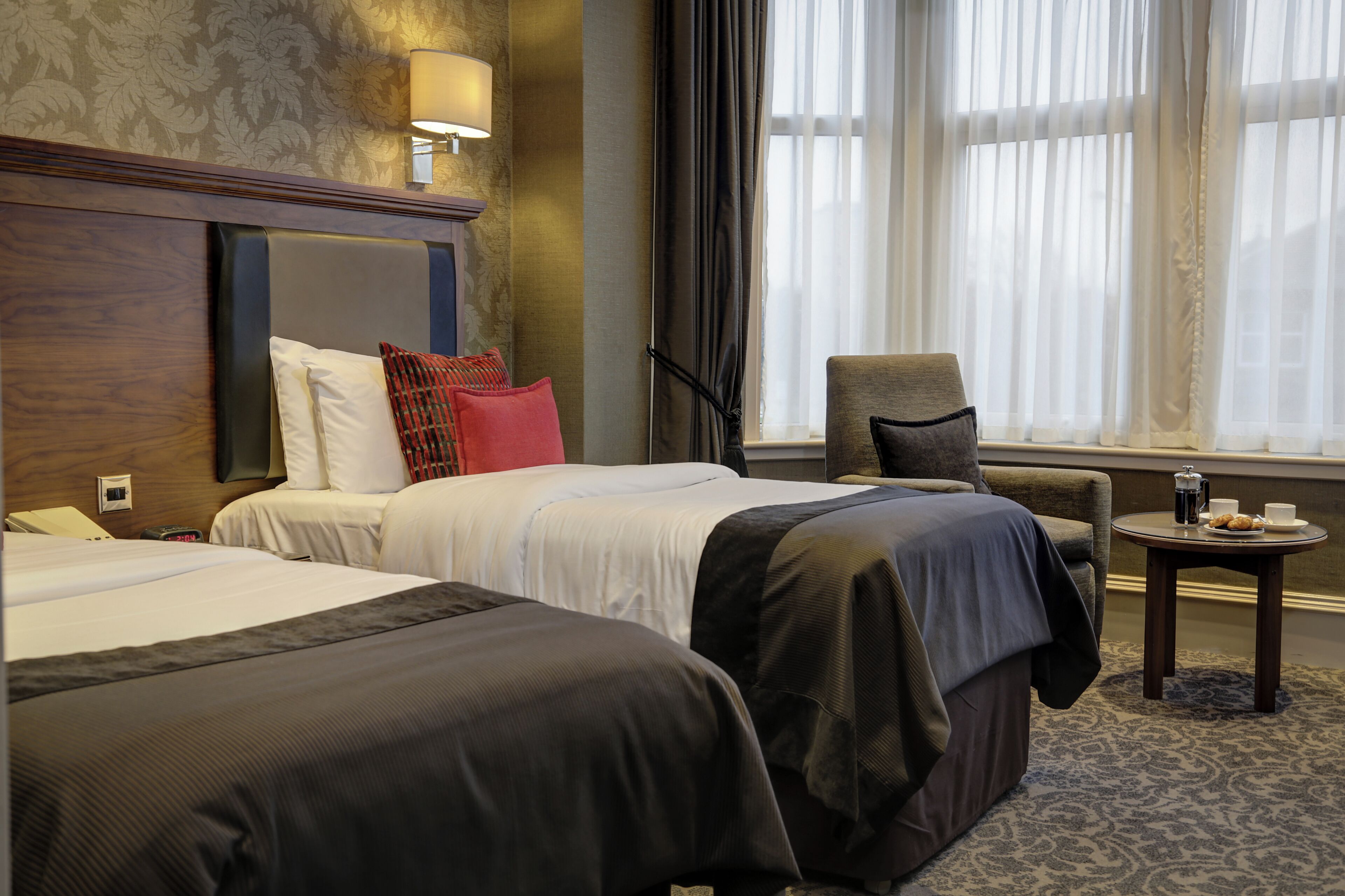A hotel room with two beds, a chair, a small table with a cup and pastries, and a window with curtains.
