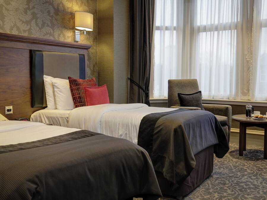 Een hotelkamer met twee bedden, een stoel, een kleine tafel met een kopje en gebak, en een raam met gordijnen.