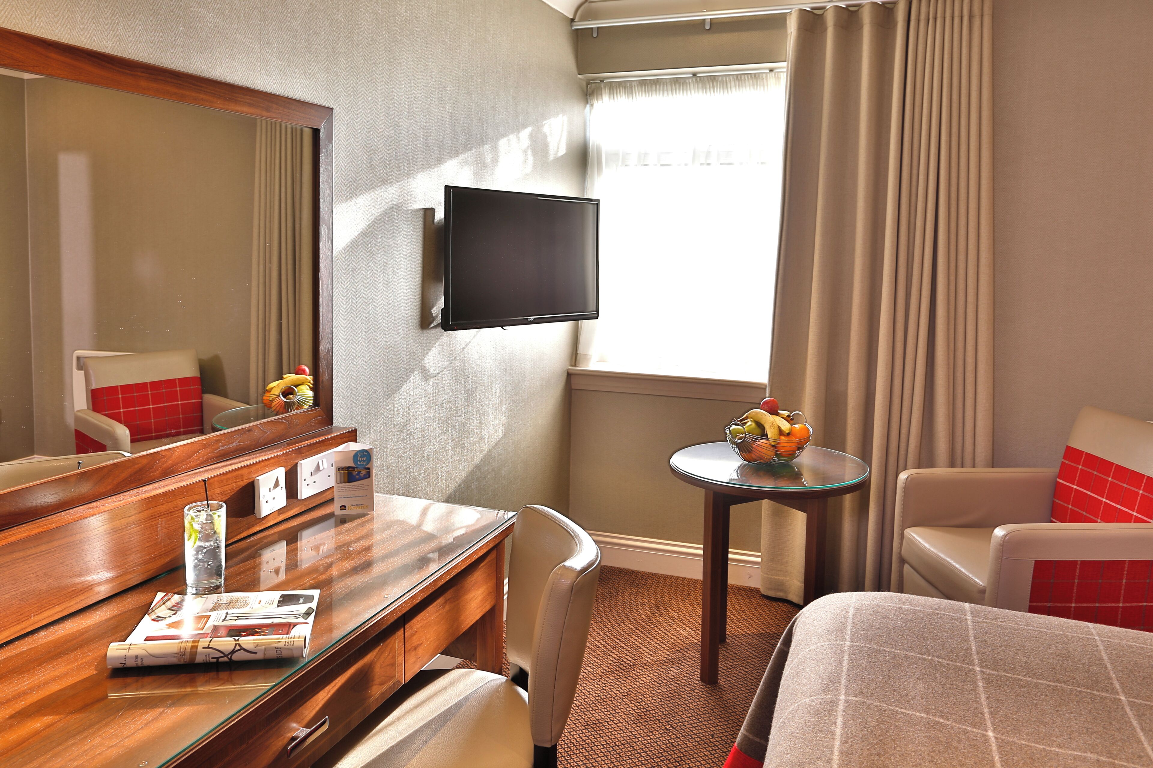 A hotel room with a bed, a desk with a chair, a small table with a fruit bowl, a TV mounted on the wall, and a window with curtains.