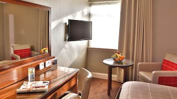 A hotel room with a bed, a desk with a chair, a small table with a fruit bowl, a TV mounted on the wall, and a window with curtains.