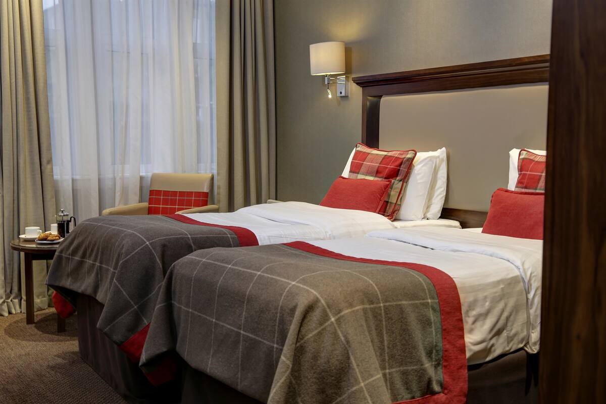 A hotel room with two beds, a nightstand, a lamp, and a window with curtains.